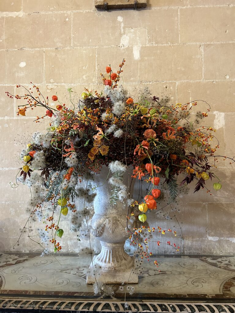 orange and green floral display in a stone urn, with ornamental grasses and dried elements. sitting on a elegant table against a stone wall in the castle Chenonceau.